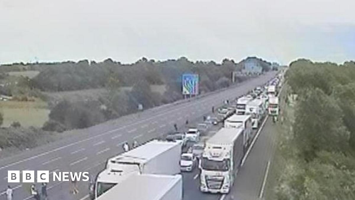 A motorway camera view showing a queue on one carriageway, People can be seen out of their cars.