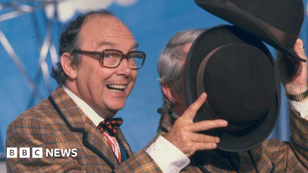 Eric Morecambe is grinning as he holds his hat to block Ernie Wise's face during a dance routine