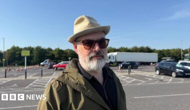 A man with a hat, sunglasses and coat standing at a car park.