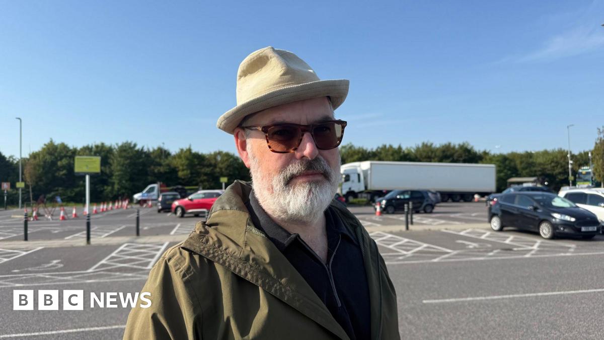 A man with a hat, sunglasses and coat standing at a car park.