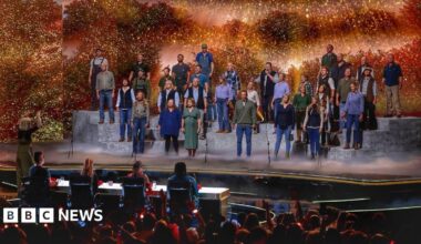A large group of performers stands on tiered platforms on a brightly lit stage, appearing to sing together as a choir. A conductor stands at the front, guiding the performance. In front of the stage, a panel of judges sits at a table, watching. The background features warm, glowing lights and foliage-like scenery, while an audience in the foreground raises hands and watches the show.