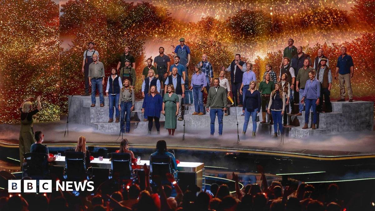 A large group of performers stands on tiered platforms on a brightly lit stage, appearing to sing together as a choir. A conductor stands at the front, guiding the performance. In front of the stage, a panel of judges sits at a table, watching. The background features warm, glowing lights and foliage-like scenery, while an audience in the foreground raises hands and watches the show.