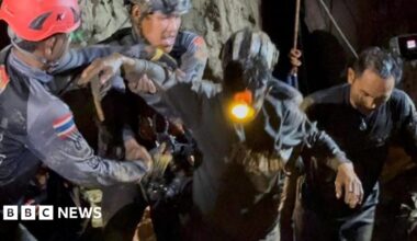 Two rescuers in dark blue diving gear, with one wearing a black helmet and the other a red one, hold the right arm of the rescued man, who is dark muddy clothes and has a headlight on. His left arm is being held by another rescuer to his left as they all emerge from the cave in darkness
