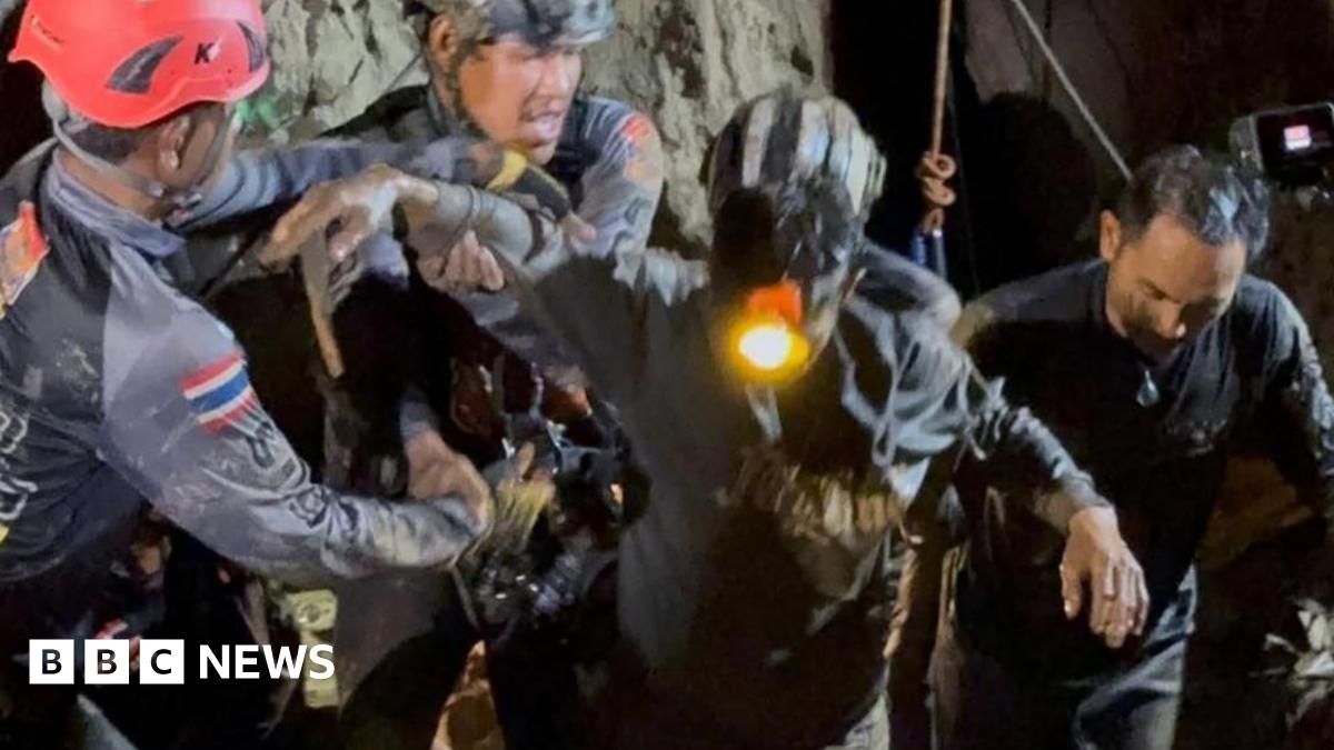 Two rescuers in dark blue diving gear, with one wearing a black helmet and the other a red one, hold the right arm of the rescued man, who is dark muddy clothes and has a headlight on. His left arm is being held by another rescuer to his left as they all emerge from the cave in darkness