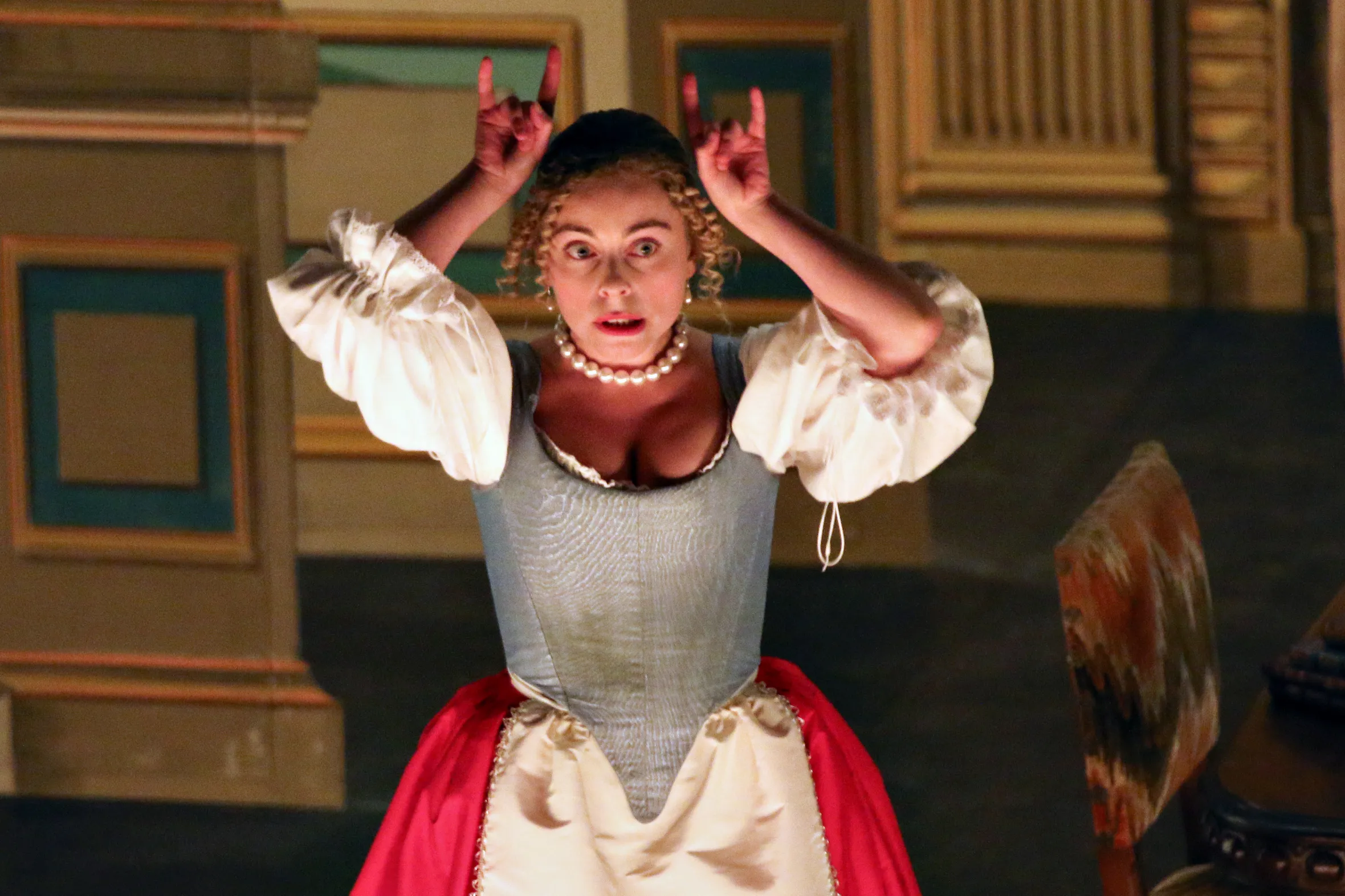 A woman dressed in period clothing with a pearl necklace, blue corset, and red skirt, with her hands raised to her head, making "horn" gestures, and a shocked expression on her face.