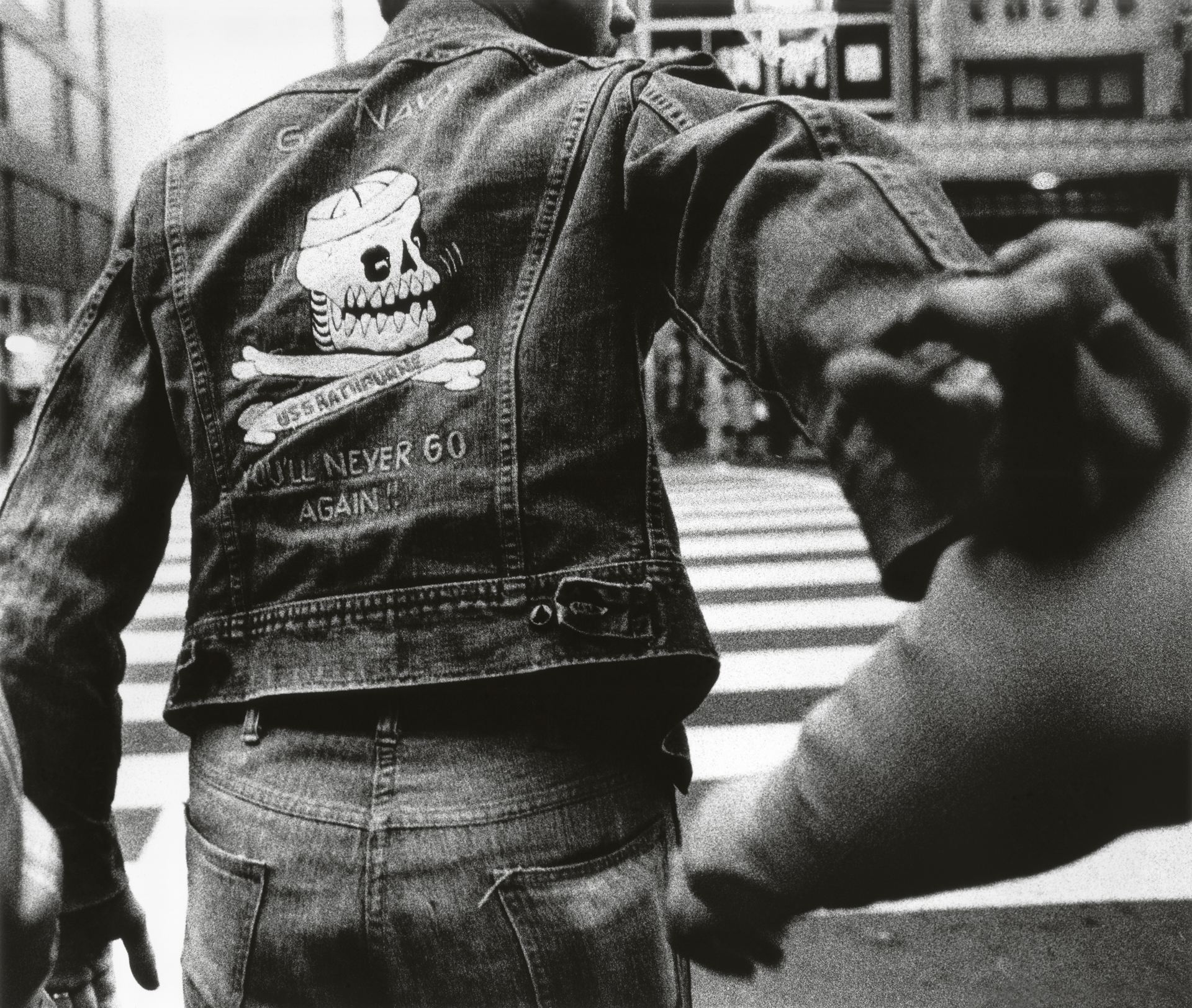 Back view of a person wearing a denim jacket with a skull and crossbones design, standing on a street.