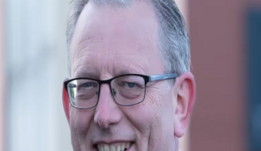 A man with short grey hair and glasses and wearing a dark blue suit, white shirt and light blue tie is smiling.