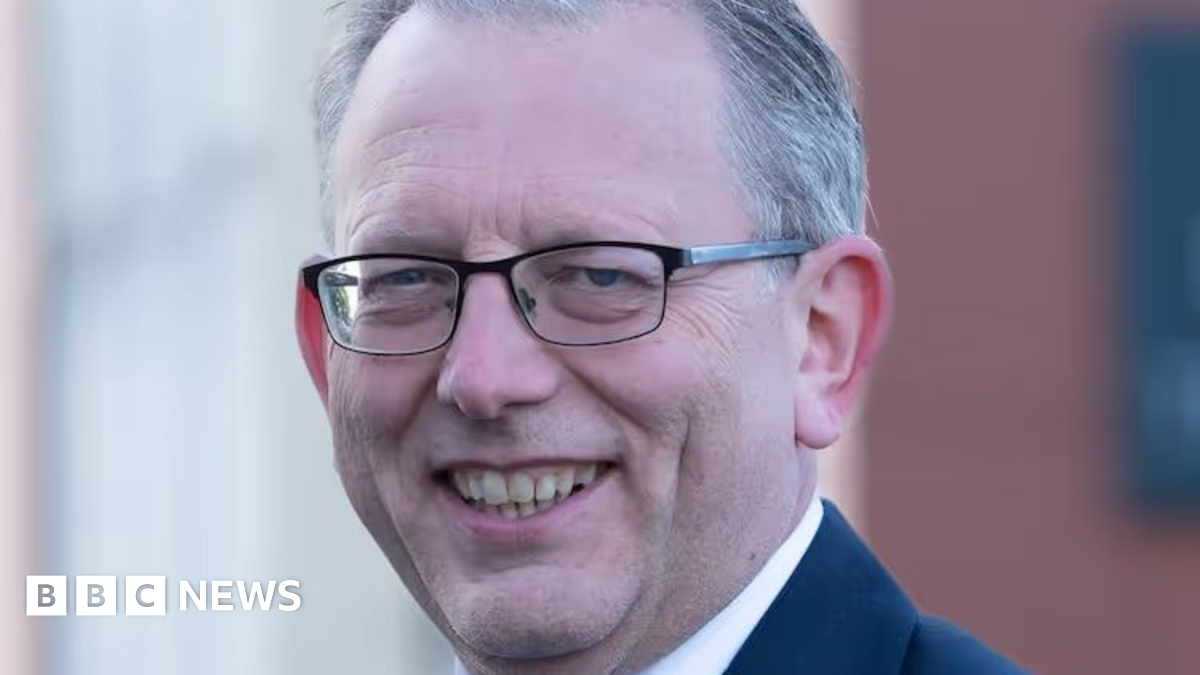 A man with short grey hair and glasses and wearing a dark blue suit, white shirt and light blue tie is smiling.