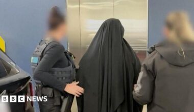 A woman wearing black Islamic dress stands in front of a lift with her back to the camera with two women in police uniform on either side