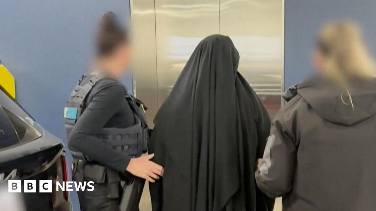 A woman wearing black Islamic dress stands in front of a lift with her back to the camera with two women in police uniform on either side