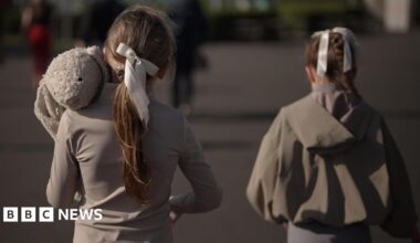 Two young girls walk alongside each other with their backs to the camera. They're wearing grey with their hair tied back in a bow. The one on the left is carrying a large stuffed toy rabbit.