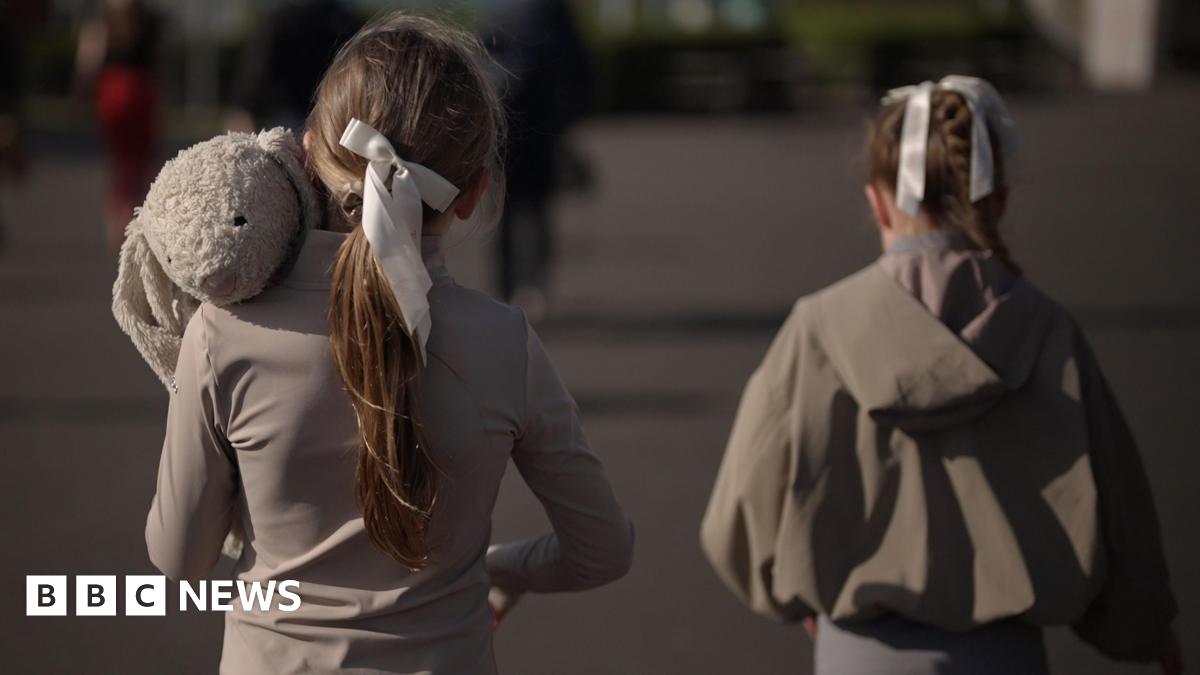 Two young girls walk alongside each other with their backs to the camera. They're wearing grey with their hair tied back in a bow. The one on the left is carrying a large stuffed toy rabbit.