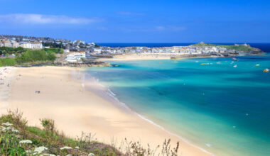 Wide view of a sunny sandy beach with turquoise water and a town of pastel houses along the coastline.