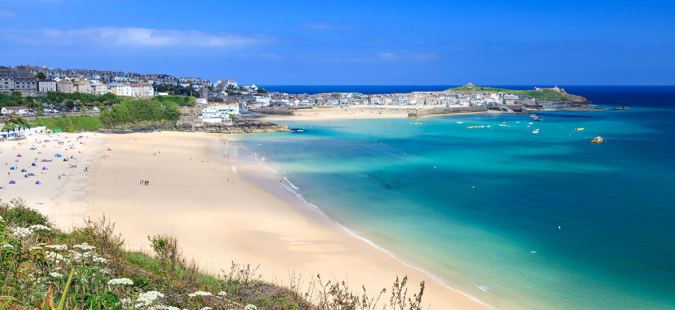 Wide view of a sunny sandy beach with turquoise water and a town of pastel houses along the coastline.