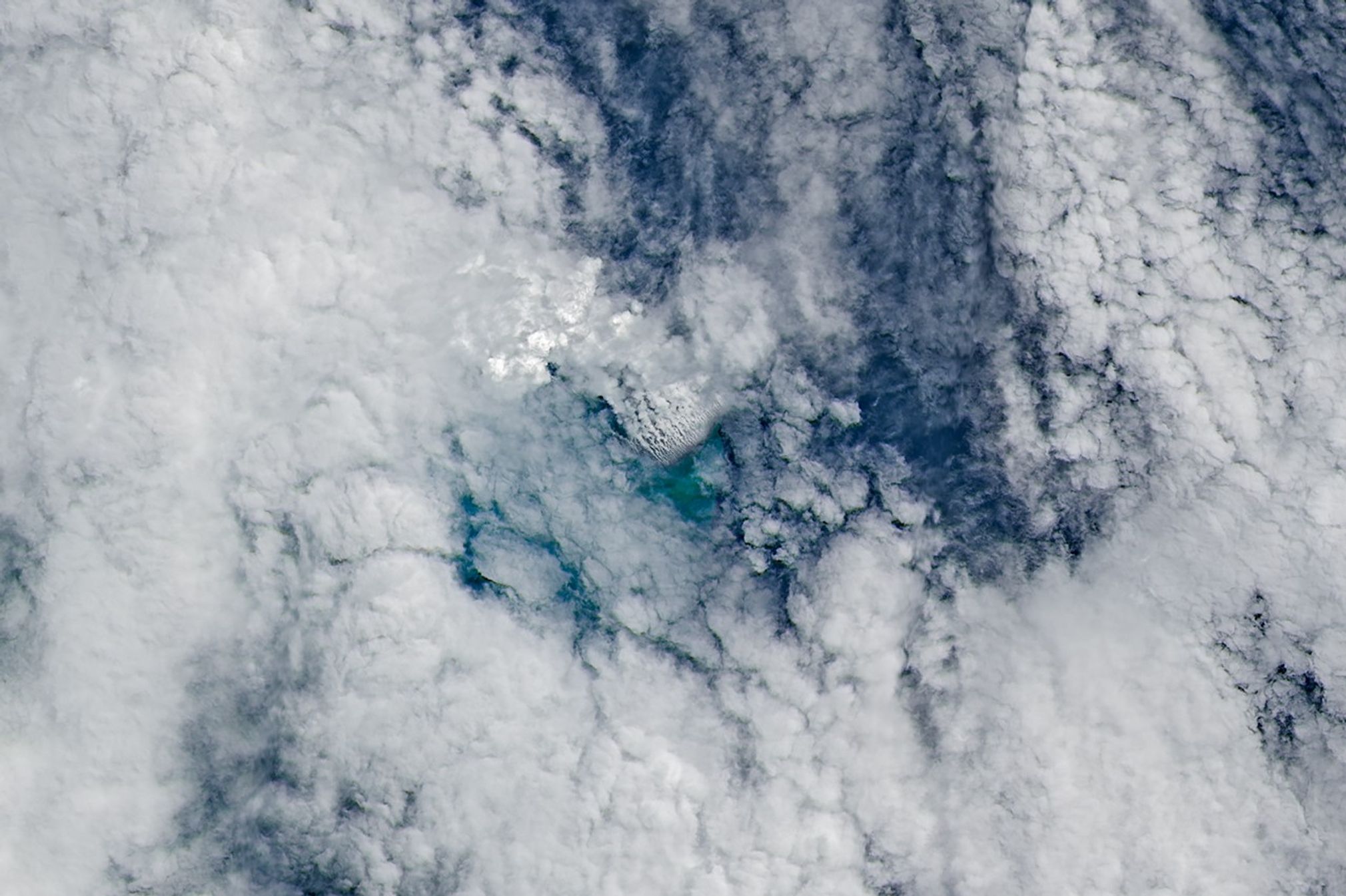 Only small patches of the ocean are visible through puffy, white volcanic plumes streaming from the center of the scene. Clouds not directly related to the eruption are visible throughout much of the image.  A false-color inset box shows the infrared signature of the eruption as a series of red dots near the volcanic plumes.