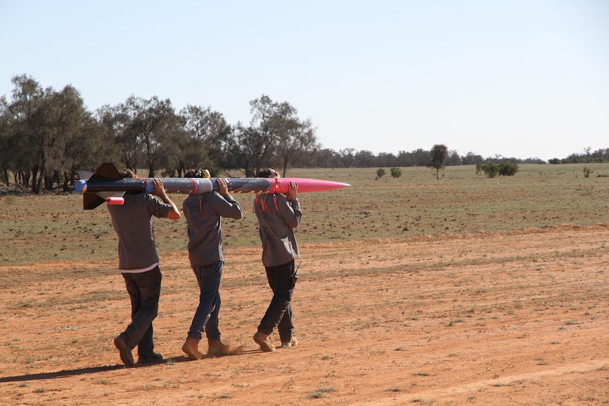 Three students walking with a grey and pink rocket on their shoulders, in a rural setting