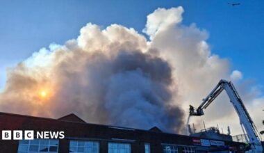 A factory building is visible with plumes of smoke billowing above it into the sky.  there is a white aerial platform with a firefighter on it, and they are spraying the fire with a hose