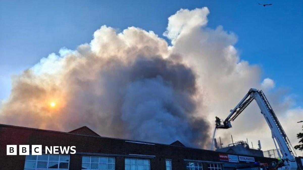A factory building is visible with plumes of smoke billowing above it into the sky.  there is a white aerial platform with a firefighter on it, and they are spraying the fire with a hose