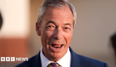 Nigel Farage has short grey hair and his mouth is open as he speaks to reporters, who are not pictured. He is wearing a blue suit jacket and a white shirt.