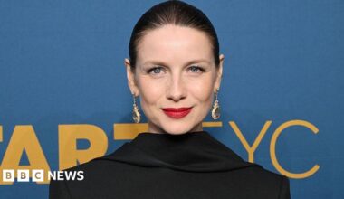 Caitriona Balfe, with her hair slicked back and wearing visible makeup including striking red lipstick as well as long jewelled earrings. She is standing in front of a blue sign with yellow writing on it.
