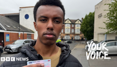 Robert is facing the camera and holding his drivers licence in his hand which he recently passed. He is standing on a residential street in Croydon with parked cars behind him.