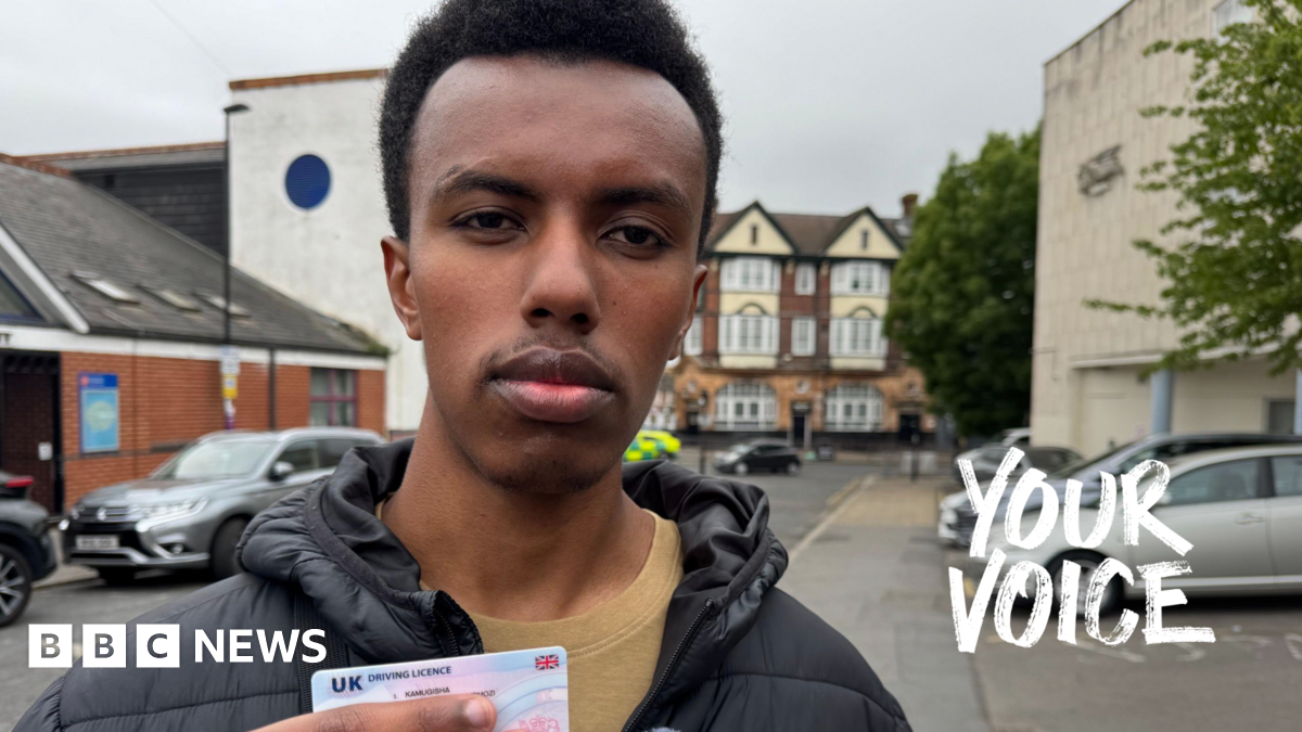 Robert is facing the camera and holding his drivers licence in his hand which he recently passed. He is standing on a residential street in Croydon with parked cars behind him.