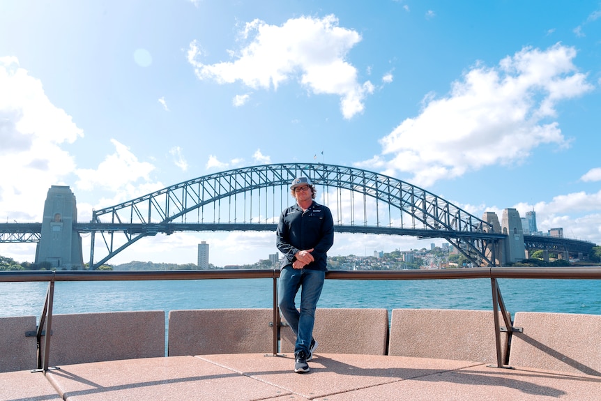 Man in jeans, navy long sleeve top and cap stands at a viewing platform by water with Sydney Harbour Bridge background.