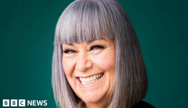 A studio portrait shows a person with a straight grey bob and fringe, wearing a black top, positioned against a plain dark green background and smiling broadly at the camera.