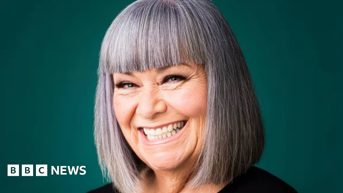 A studio portrait shows a person with a straight grey bob and fringe, wearing a black top, positioned against a plain dark green background and smiling broadly at the camera.