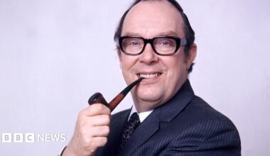 Head and shoulders of Eric Morecambe in1973. He is sideways on to the camera smiling with a pipe in his right hand the end of which is between his teeth. He is wearing a fine pinstripe dark blue suit, white shirt and patterned tie, has receding dark hair and dark-framed glasses