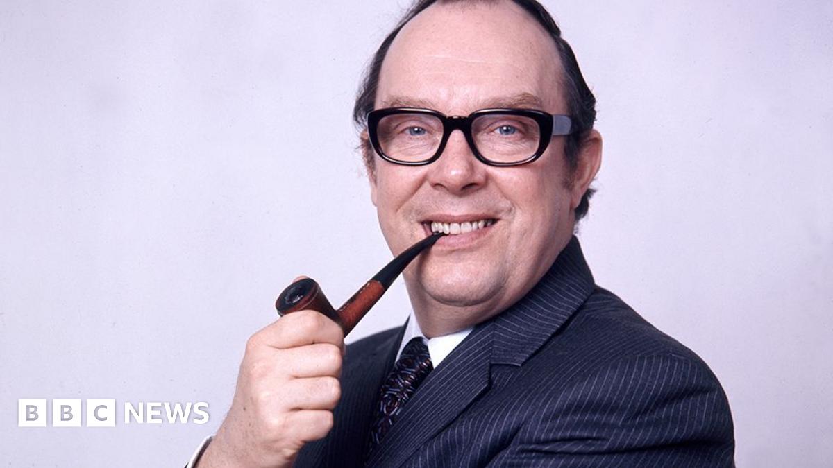 Head and shoulders of Eric Morecambe in1973. He is sideways on to the camera smiling with a pipe in his right hand the end of which is between his teeth. He is wearing a fine pinstripe dark blue suit, white shirt and patterned tie, has receding dark hair and dark-framed glasses