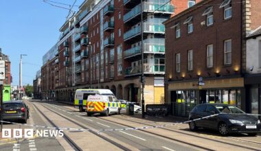 Blue and white police tape stretches across a city centre street. behind the tape, two police vans and a police car are parked , blocking the junction to a side street. The bar is a three-storey red brick building with floor to ceiling windows at ground level and two rows of four standard windows on the floors above. The next building along the street is a modern seven-storey block of apartments with glass-fronted balconies. It is a sunny day with a blue sky.
