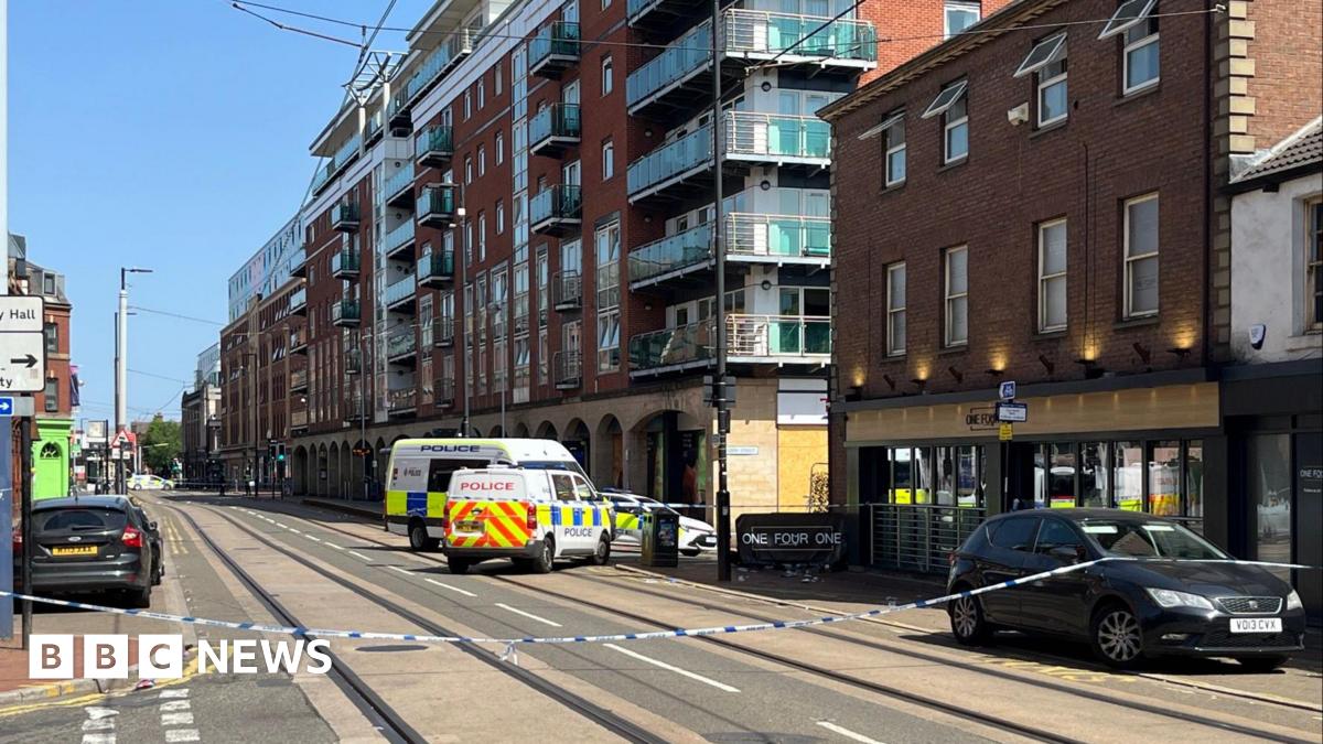 Blue and white police tape stretches across a city centre street. behind the tape, two police vans and a police car are parked , blocking the junction to a side street. The bar is a three-storey red brick building with floor to ceiling windows at ground level and two rows of four standard windows on the floors above. The next building along the street is a modern seven-storey block of apartments with glass-fronted balconies. It is a sunny day with a blue sky.