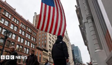 A street in the US with pedestrians walking around and a US flag hanging from a building.