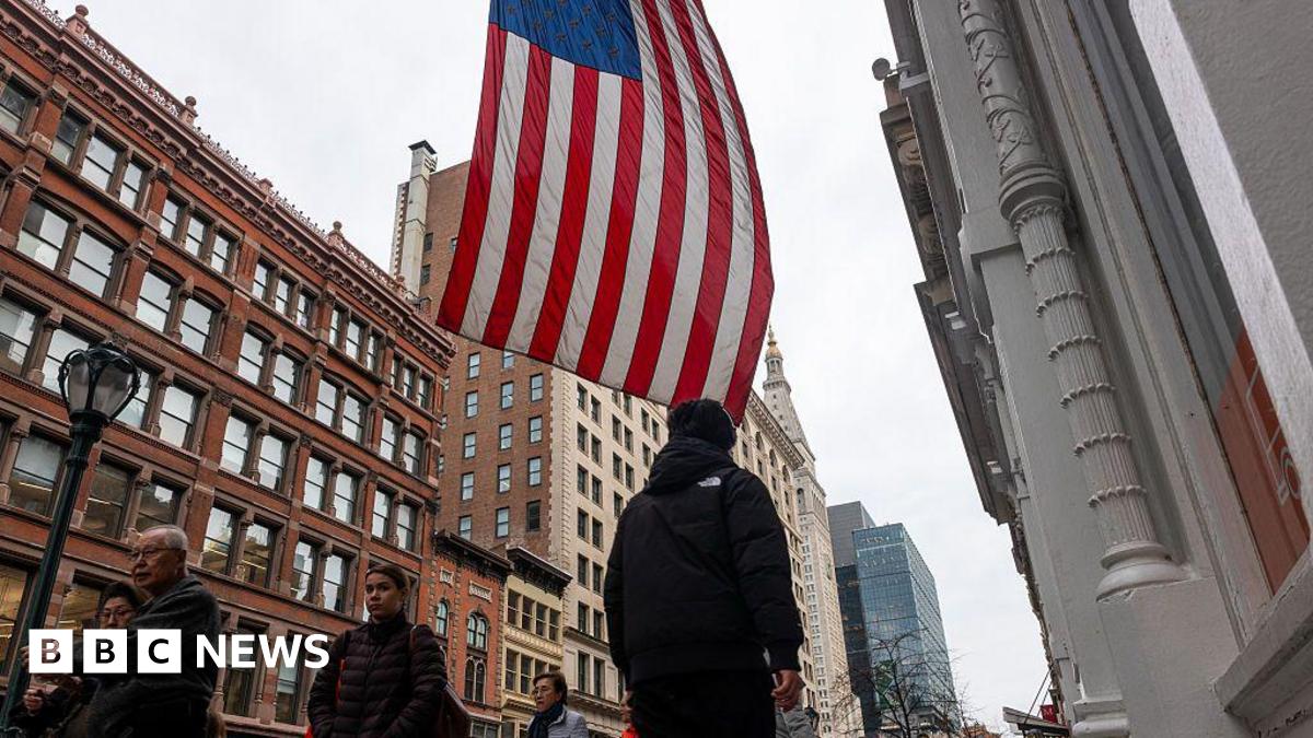 A street in the US with pedestrians walking around and a US flag hanging from a building.