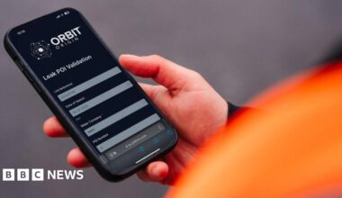 A person's zoomed-in hand holding a black smartphone which displays an app.  The apps title is Orbit Origin.  There is a blurred orange jacket to the left of the picture along with a blurred grey background.