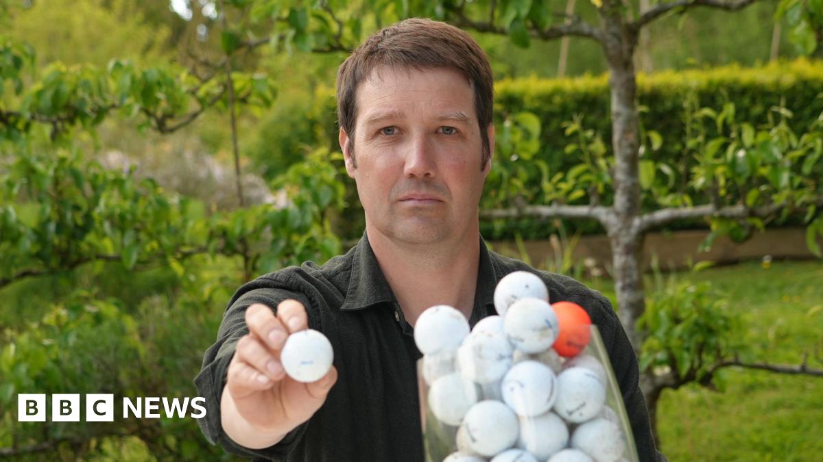 A head and shoulders image of a man wearing a dark coloured shirt while sitting in a garden with trees behind him. He is holding a white golf ball to the camera while looking into the camera. In front of him is a clear receptacle filled with golf balls - all white ones, except for one orange one.