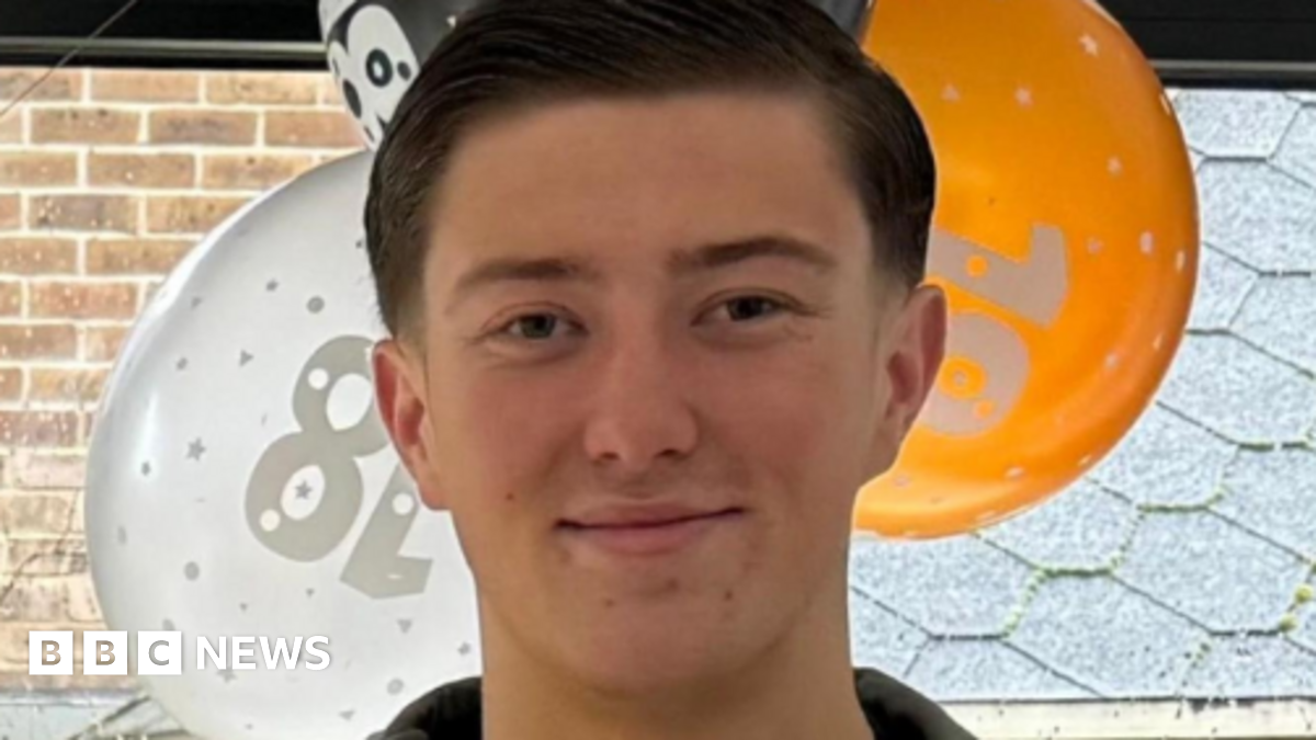 An 18-year-old man standing in front of orange, white and black balloons by a window. He has short dark hair and is smiling and is wearing a blue jacket and a black top underneath.