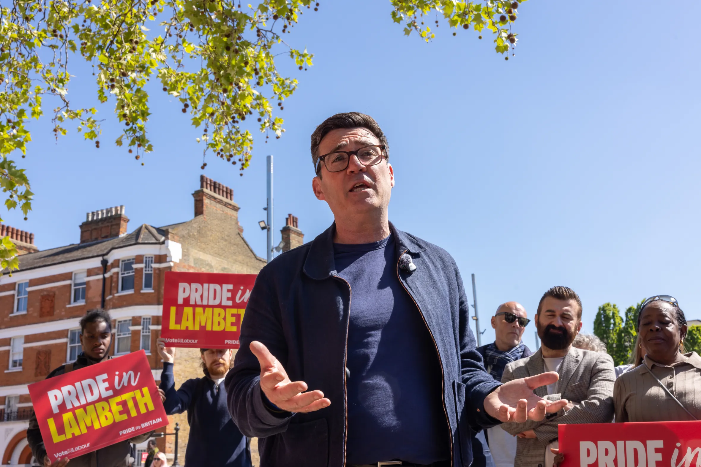 Andy Burnham speaking in Brixton, London, with people holding "Pride in Lambeth" signs in the background.