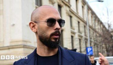 A close-up shot of Andrew Tate, wearing a dark blue suit with a black top underneath, from the shoulders up. He has a bald head, sunglasses, and a dark beard.