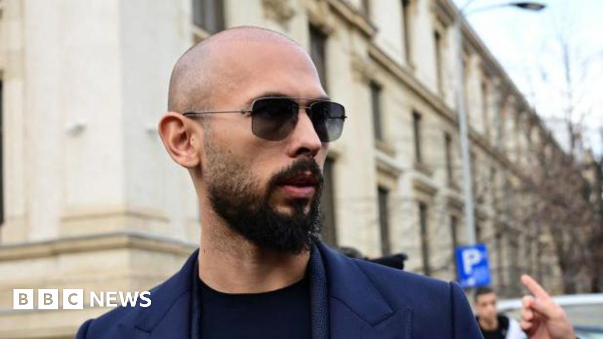 A close-up shot of Andrew Tate, wearing a dark blue suit with a black top underneath, from the shoulders up. He has a bald head, sunglasses, and a dark beard.