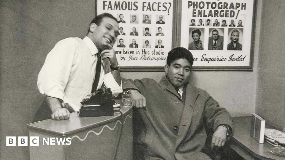 Dave Sharkey, with customer, 1962. Black‑and‑white photograph of Dave Sharkey, with customer. Dave leans against a counter with a camera placed on top, while the other sits beside the desk. Posters of sample portrait photographs hang on the wall behind them.