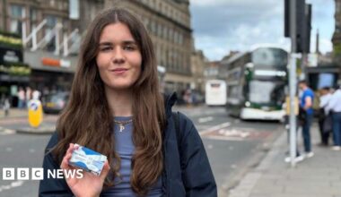 Poppy Fairbairn holds the National Entitlement Card towards the camera with a blurred Lothian Bus in the background.