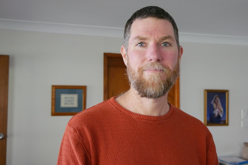 A man with a beard standing behind a wall with pictures hang in the background.