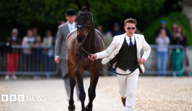 A smart young man, in a white suit with a black waistcoat and blue tie, running along a path whilst holding the reins of a a race horse. There is a man in a grey suit and bowler hat behind him and railings with spectators standing in the background
