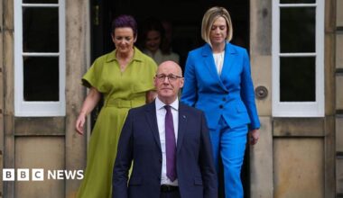 John Swinney wearing a dark suit and purple tie standing on the steps of Bute House. Behind him, members of his cabinet are emerging from a door.