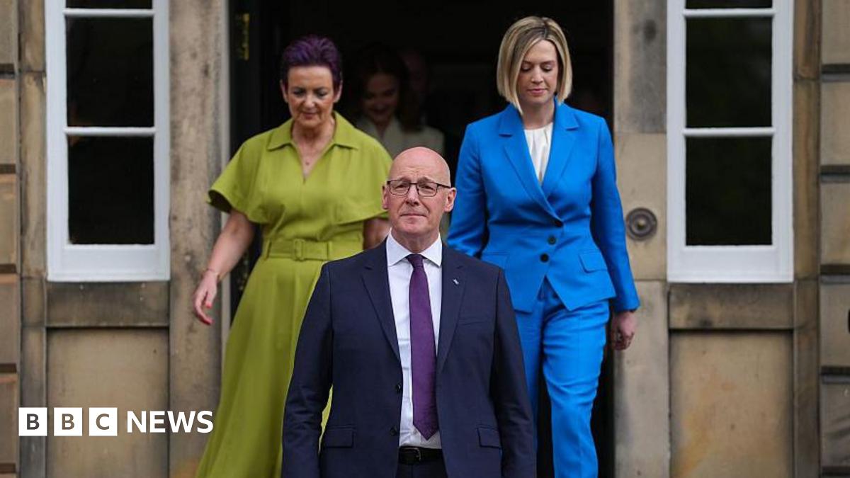 John Swinney wearing a dark suit and purple tie standing on the steps of Bute House. Behind him, members of his cabinet are emerging from a door.