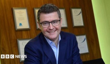 Brendan Mooney smiles while sitting at a desk with a Kainos sign above his head. He has short dark hair and black-framed glasses and is wearing a navy suit jacket and open-necked shirt