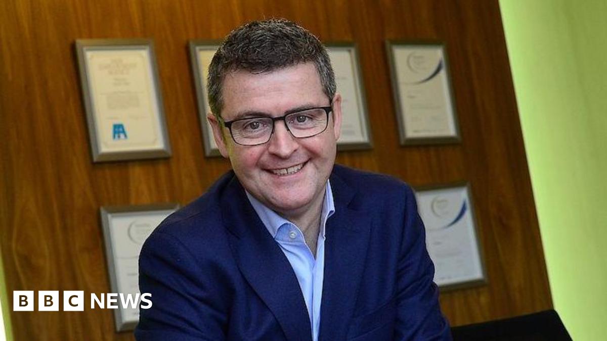 Brendan Mooney smiles while sitting at a desk with a Kainos sign above his head. He has short dark hair and black-framed glasses and is wearing a navy suit jacket and open-necked shirt