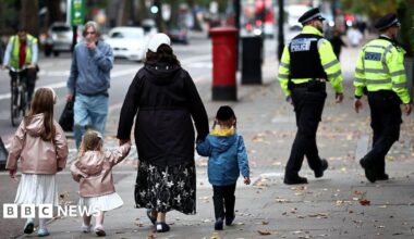 A family including two younger girls in white dresses and pink coats along with a little boy in a blue coat. They walk down the street with their backs to the camera and in the background two Metropolitan Police officers in uniform can be seen walking in the same direction.
